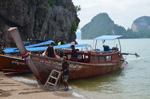 Phang Nga, Thailand - December 2, 2018: Local people prepare tourist and fishing boats for sailing. Fancy boats at the shore in the bay on the background of green mountains.