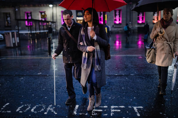 Visually impaired man walking with the assistance of a woman through City Centre, he is using a white walking cane for guidance as well as holding his friends arm. Its dark and raining as they cross the road.