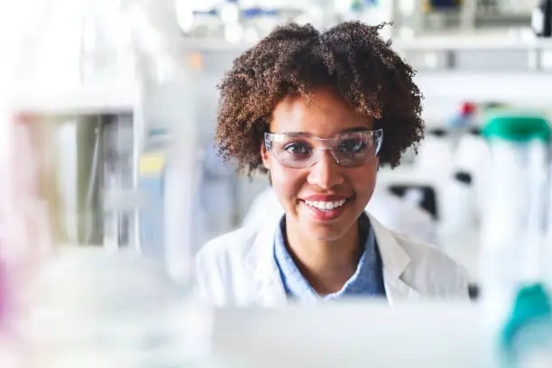 Close-up portrait of young scientist smiling. Confident female researcher is wearing protective eyewear. She is in science laboratory.