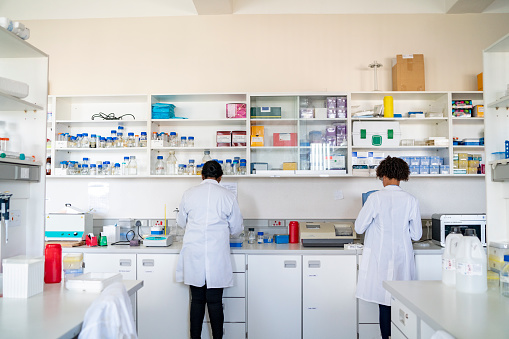 Rear view of female scientists working in laboratory. Multi-ethnic researchers are doing scientific experiment. They are wearing lab coats.