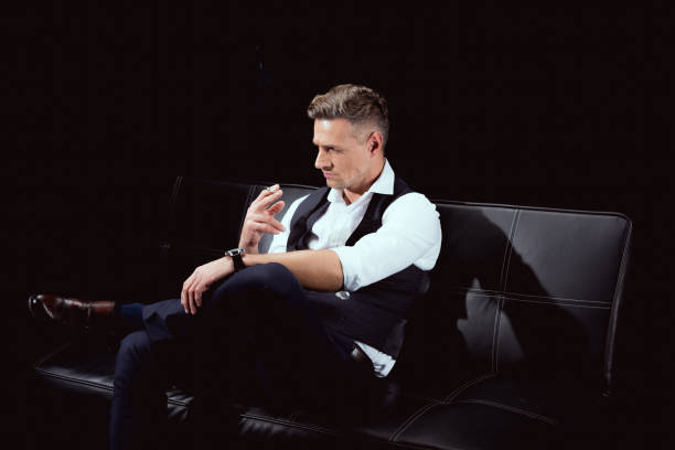 istock handsome man in formal wear sitting on couch with cigarette isolated on black 1094850150