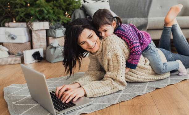 innenaufnahme der schöne glückliche junge frau am laptop in gemütlichen innenraum weihnachten online-shopping. mutter auf dem boden liegend umarmen als nächstes den weihnachtsbaum und sofa und tochter, geschenke einkaufen. - christmas shopping internet family stock-fotos und bilder