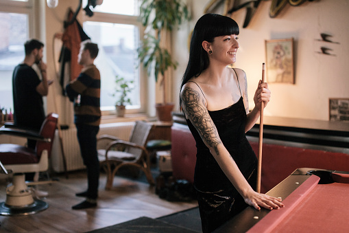 Tattooed woman standing at Billiard Table smiling. Male friends are standing in the background.