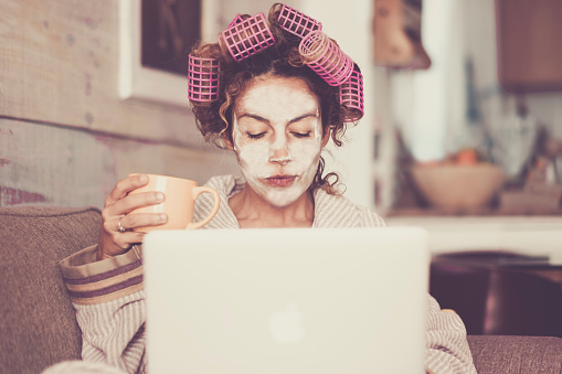 Rare and funny portrait of beautiful woman with facial mask and curlers hair taking a coffee at home while use internet with modern laptop computer - home scene for modern lifestyle people taking his own time