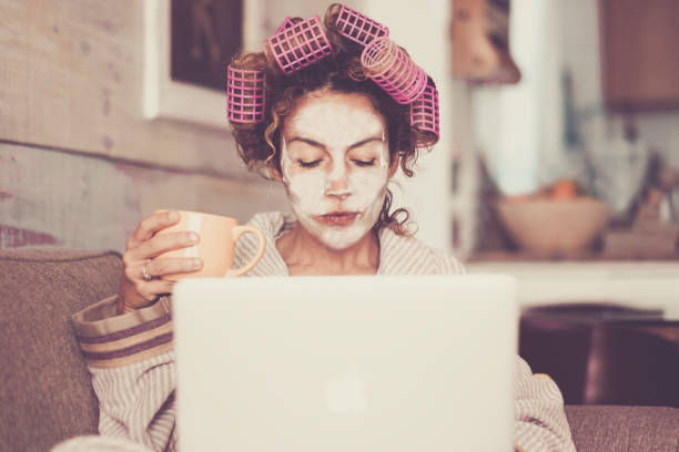 rare and funny portrait of beautiful woman with facial mask and curlers hair taking a coffee at home while use internet with modern laptop computer - home scene for modern lifestyle people taking his own time - hairstyle human hair women retro revival imagens e fotografias de stock