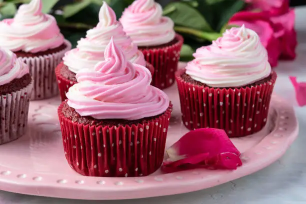Close up of pink and white swirled buttercream frosting on a red velvet cupcake. Cupcakes are on a decorative pink plate. Fresh pink rose petals adorn the table and fresh pink roses are seen in the background.