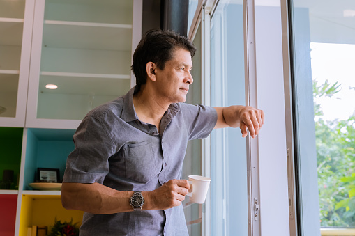 Middle-aged Asian man looking through a window, sipping coffee and using ideas