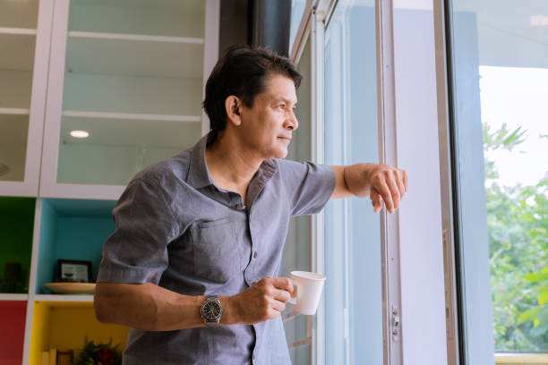 azjatycki mężczyzna w średnim wieku patrzy przez okno, popija kawę i korzysta z pomysłów - men thinking contemplation pensive zdjęcia i obrazy z banku zdjęć