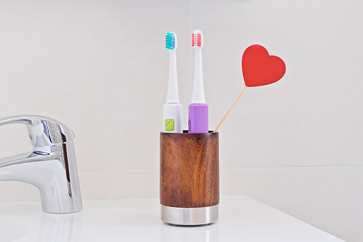 Two electric toothbrushes in wooden glass on sink in bathroom with red heart. Toothbrush for personal healthcare dental care. Oral dental hygiene. Dental equipment. Morning routine concept