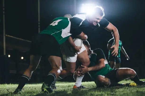 Strong rugby player trying to escape the tackle from opposite team. Rugby team players competing in game under lights.
