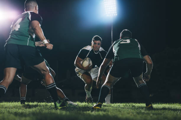 Rugby players tackling during game Rugby player running on field with ball and tackling with opposite team players during the game. Professional rugby action under lights at sports arena. rugby team stock pictures, royalty-free photos & images