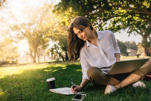 Businesswoman sitting on grass at park with laptop and making notes in her diary. Freelancer female working while sitting at park.