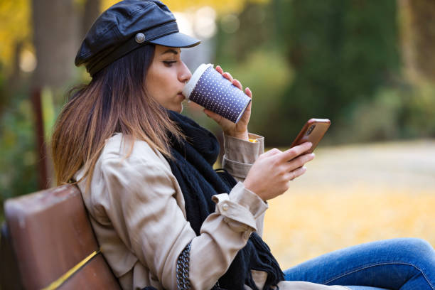 istock Beautiful young woman using her mobile phone while drinking coffee and sitting in a bench in the street. 1095043684