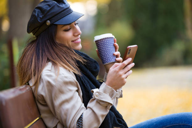 istock Beautiful young woman using her mobile phone while drinking coffee and sitting in a bench in the street. 1095043720