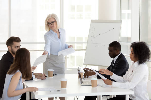 Smiling mature manager giving handouts to diverse employees at meeting Smiling mature female manager company leader gives handout to diverse employees team at office group meeting, middle aged businesswoman handing paper finanical report to workers at corporate briefing delegating stock pictures, royalty-free photos & images