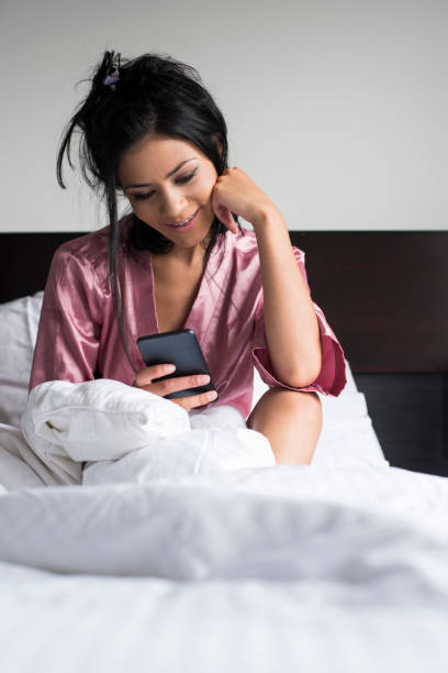 Woman lying down watching cell phone Woman lying on her bed, wearing her pink pajamas, watching her cell phone, playing with him and taking a selfie, in the comfort of her room tecnología inalámbrica stock pictures, royalty-free photos & images