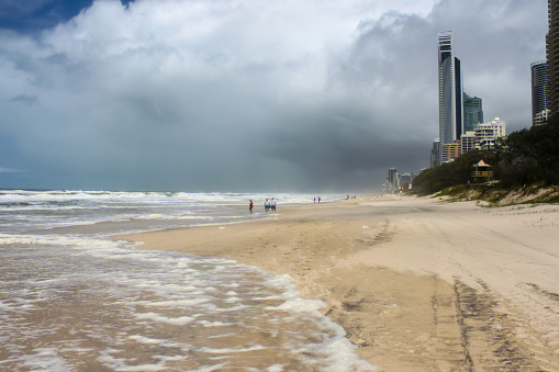 Tire tracks and unidentifiable tourists on beach wading out into surf on stormy day with dark clouds at Gold Coast - Surfers Paradise Australia with city in background