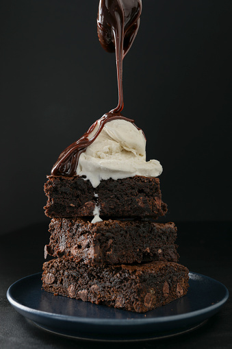 An extreme close up vertical photograph of hot fudge being poured over a stack of freshly baked whole wheat  brownies.Isolated on black.