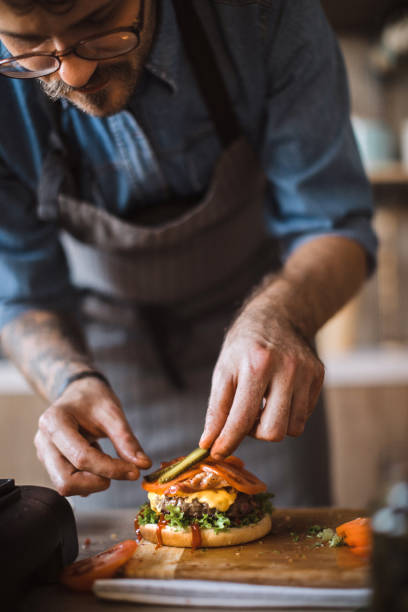 Adding Final Details to Delicious Cheeseburgers Young Male Cook Adding Final Details to Delicious Cheeseburgers food state stock pictures, royalty-free photos & images