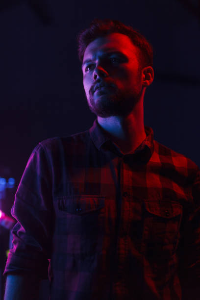 Handsome bearded hipster in shirt standing in dark glowing neon lights looking away.