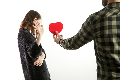 Portrait of a young couple having conflict. Man asking for forgiveness offering a red heart-shaped gift to his depressed girlfriend