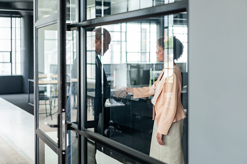 Smiling businesswoman shaking hands with businessman. Colleagues are greeting at new office. They are seen through glass wall.