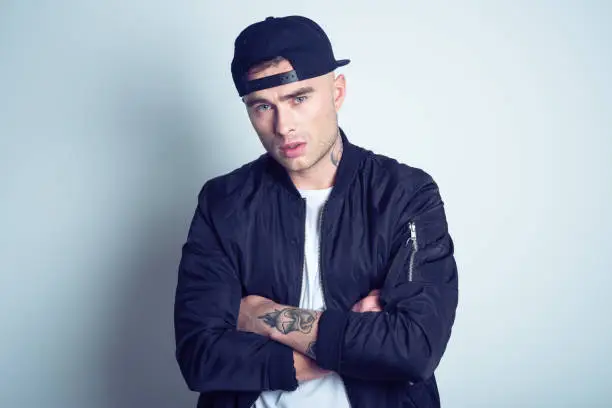 Studio portrait of angry, tattooed young man wearing black bomber jacket and baseball cap, staring at camera.