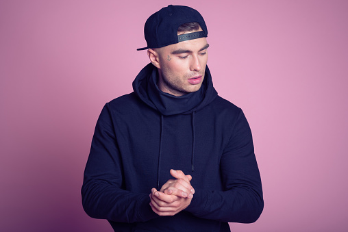 Studio portrait of tattooed young man wearing black hooded shirt and baseball cap, standing against pink background.