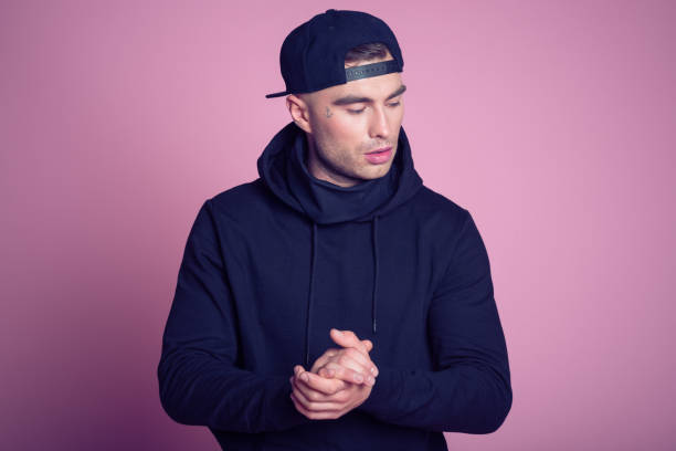 Portrait of rebellious young man wearing hooded shirt Studio portrait of tattooed young man wearing black hooded shirt and baseball cap, standing against pink background. rap stock pictures, royalty-free photos & images