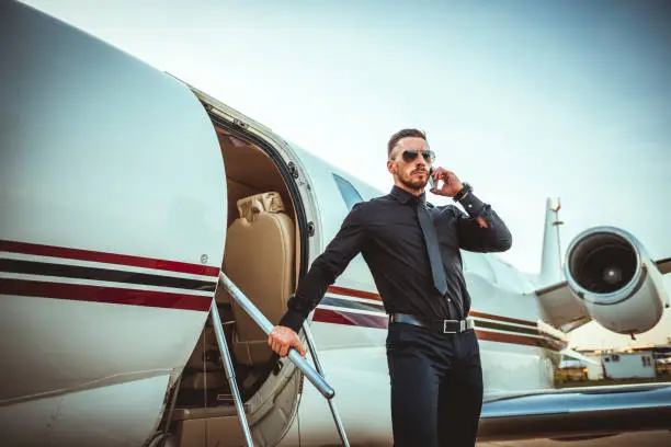 Young rich man disembarking from a private jet. He is having a conversation on a smart phone.