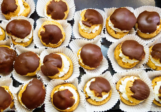 Looking down on a large group of eclairs, or choux buns, filled with cream and covered in chocolate frosting, each resting in a paper case. Sweet temptation!
