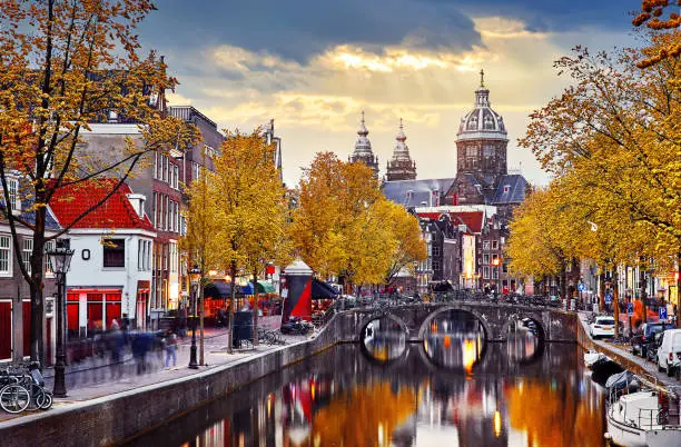 Amsterdam, Netherlands. Autumn sunset in Red-light district. View at Church saint Nicholas above bridge canal with boats. Embankment street lamp and yellow trees Evening streets landscape.