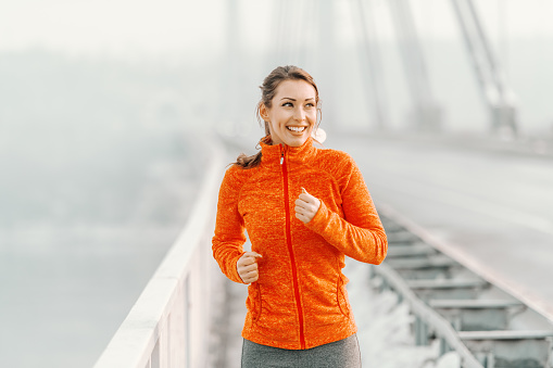 Happy Caucasian woman in sportswear and with ponytail running on the bridge at winter. Winter fitness concept.