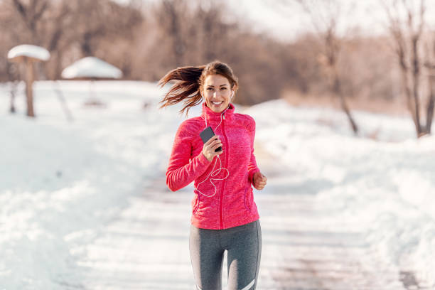 femme de race blanche heureuse souriante sportswear debout sur la piste avec écouteurs sur et écouter de la musique. concept de remise en forme d’hiver. - beautiful caucasian teenager running photos et images de collection