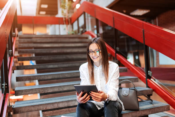 étudiante souriante avec les lunettes et les cheveux bruns avec tablette en position assise sur les marches. à côté de son sac. - women computer home interior brown hair photos et images de collection