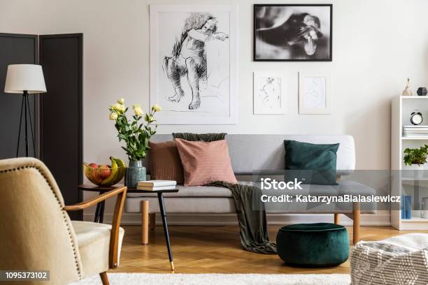 Flowers On Wooden Table In Front Of Settee Under Posters In Flat Interior With Armchair Real Photo Stock Photo - Download Image Now