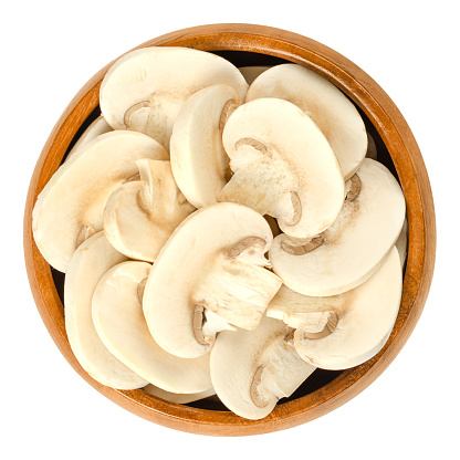 Sliced white champignon mushrooms in wooden bowl. Agaricus bisporus, also called common, button, cultivated or table mushroom. Isolated macro food photo closeup from above on white background.