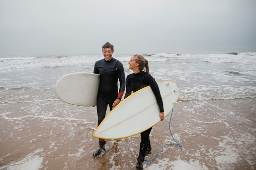 Young couple walking back onto the shore after surfing in the North Sea. They are holding their surfboards, smiling, laughing and talking.