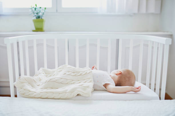 Adorable baby girl sleeping in co-sleeper crib attached to parents' bed. Little child having a day nap in cot. Sleep training concept. Infant kid in sunny nursery