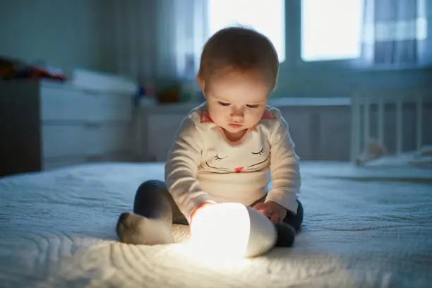 Photo of Adorable baby girl playing with bedside lamp in nursery
