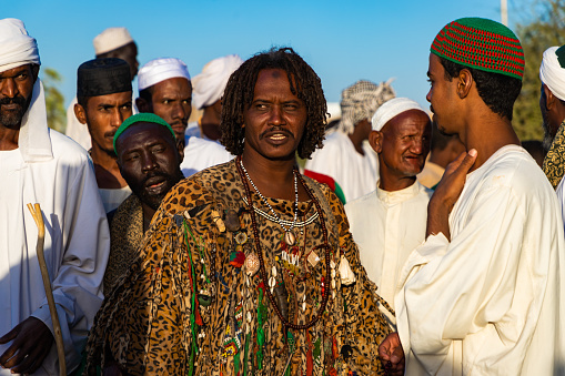 Omdurman, Sudan – December 7, 2018: Highlight of visiting Sudan would be this Sufis ritual as well as Pyramids. Sufis gather every Friday at Hamed al-Nil Tomb in Omdurman, which is a short drive from capital of Sudan, Khartoum. Tons of Sufis start marching around sunset with sticks and flags respectively. As far as I could see, it was only men. Tourist can go watch this ceremony with no issue. After they make a huge circle in front of the mosque, ceremony finally starts. As their chant grows louder, everybody starts moving his body to the chanting happily and clap. They are not in trance but just enjoy the ceremony. Some lead the ceremony with loud voice in the center of the circle, others swirl. Most dervishes wear white thobe but some wear very outstanding costume, which are very colourful and unique patchwork gown or animal printed robe. Surprisingly those who are in outstanding costume have dreadlocks hair !
