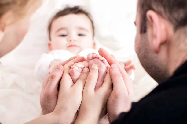 The image captures a tender moment between an adult and a baby, focusing on their hands. The adult's hand, weathered and strong, gently holds the tiny, delicate hand of the baby. The contrast between the rough texture and larger size of the adult's hand and the softness and fragility of the baby's fingers highlights the themes of care, protection, and love. The baby is dressed in a soft, knitted white garment, adding a sense of warmth and purity to the scene. The background is softly blurred, ensuring that the viewers attention is drawn entirely to the touching connection between the two hands, symbolizing a bond that is both nurturing and profound.