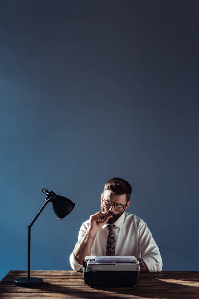 istock handsome journalist sitting at table with lamp and retro typewriter and smoking on grey background 1095495918
