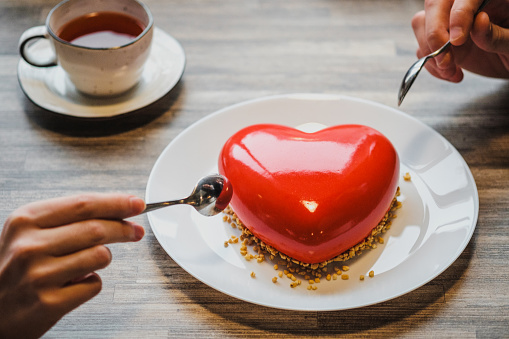 Red cake in the shape of a heart is on the table. Two hands with spoons, male and female, stretch into a pie. Romantic date, Valentine's Day.