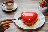 Red cake in the shape of a heart is on the table. Two hands with spoons, male and female, stretch into a pie.