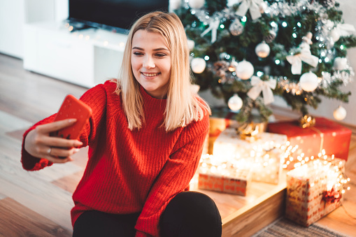 Young blonde woman sitting next to the Christmas tree, taking a photo, selfie with her phone. Wearing a red christmas sweater.