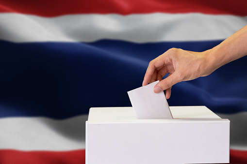 Close-up of human hand casting and inserting a vote and choosing and making a decision what he wants in polling box with Thailand flag blended in background