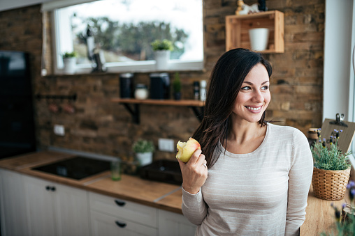 Portrait of a smiling woman eating apple and looking through window while standing in the kitchen.