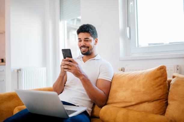 sorrindo casual freelancer de raça mista usando telefone inteligente e portátil enquanto está sentado no sofá. - web browsing - fotografias e filmes do acervo