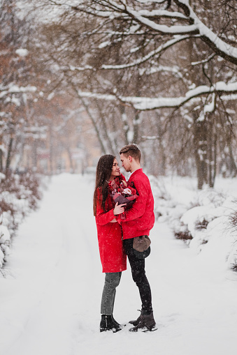A date of lovers with my park in the winter. A bouquet of red flowers, walk, hug, kiss, laugh in a romantic setting. Portrait of a couple, husband and wife. Red jackets and coats. St. Valentine's Day.